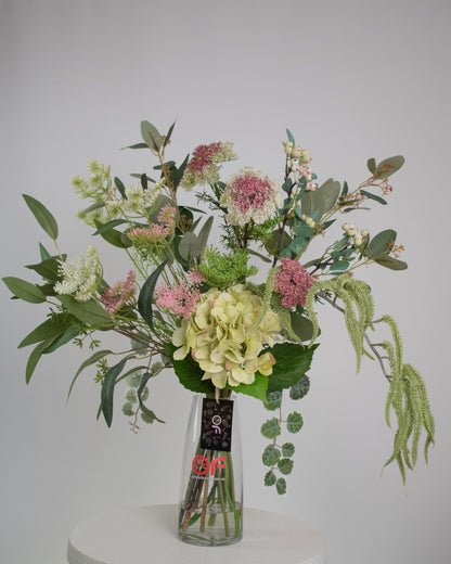 Artificial floral arrangement featuring green hydrangeas, pink wildflowers, and lush foliage in a clear glass vase with acrylic water