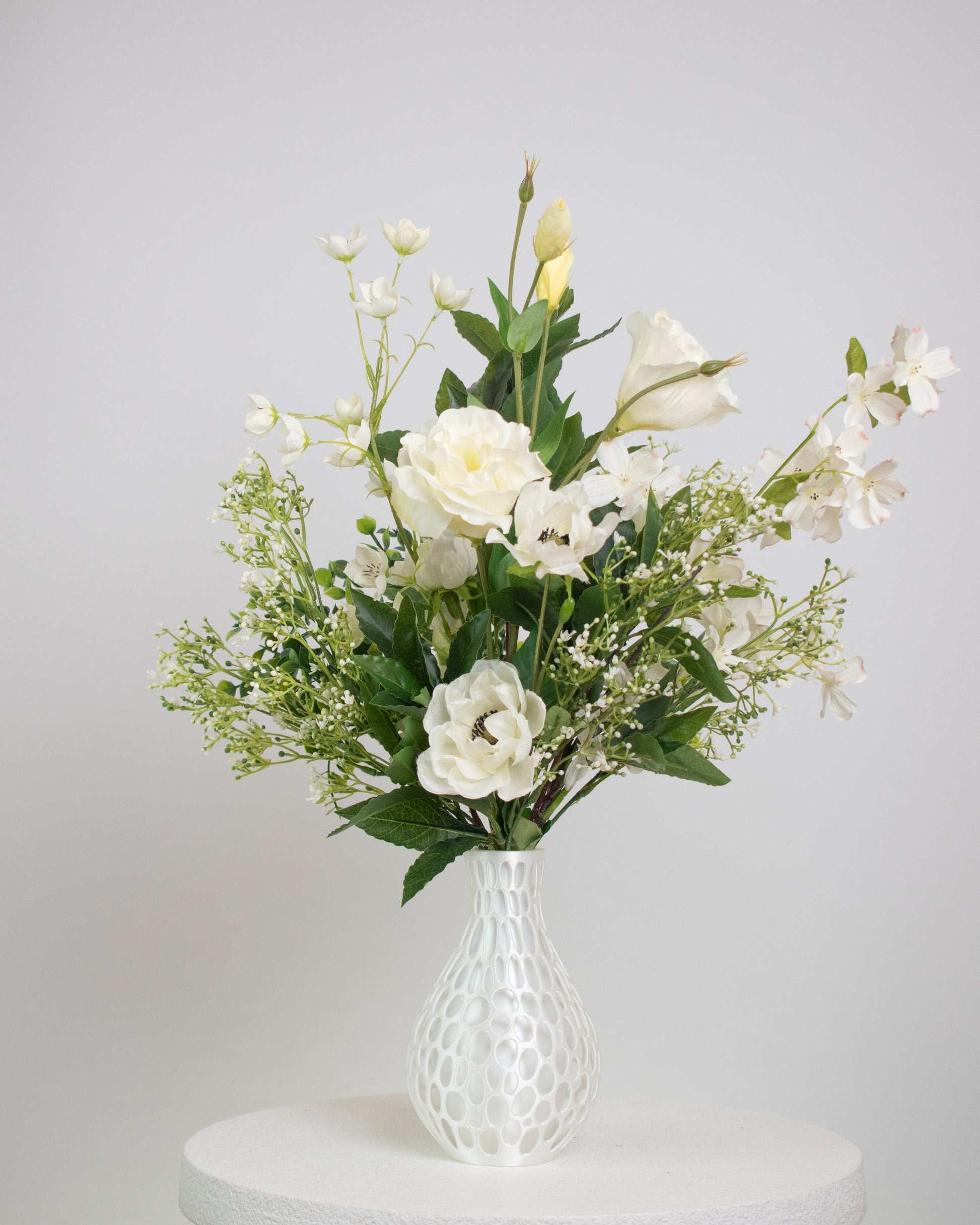 Artificial floral arrangement featuring ivory roses, white lisianthus, and blossoms with lush green foliage, styled in a white honeycomb-textured vase on a neutral background.