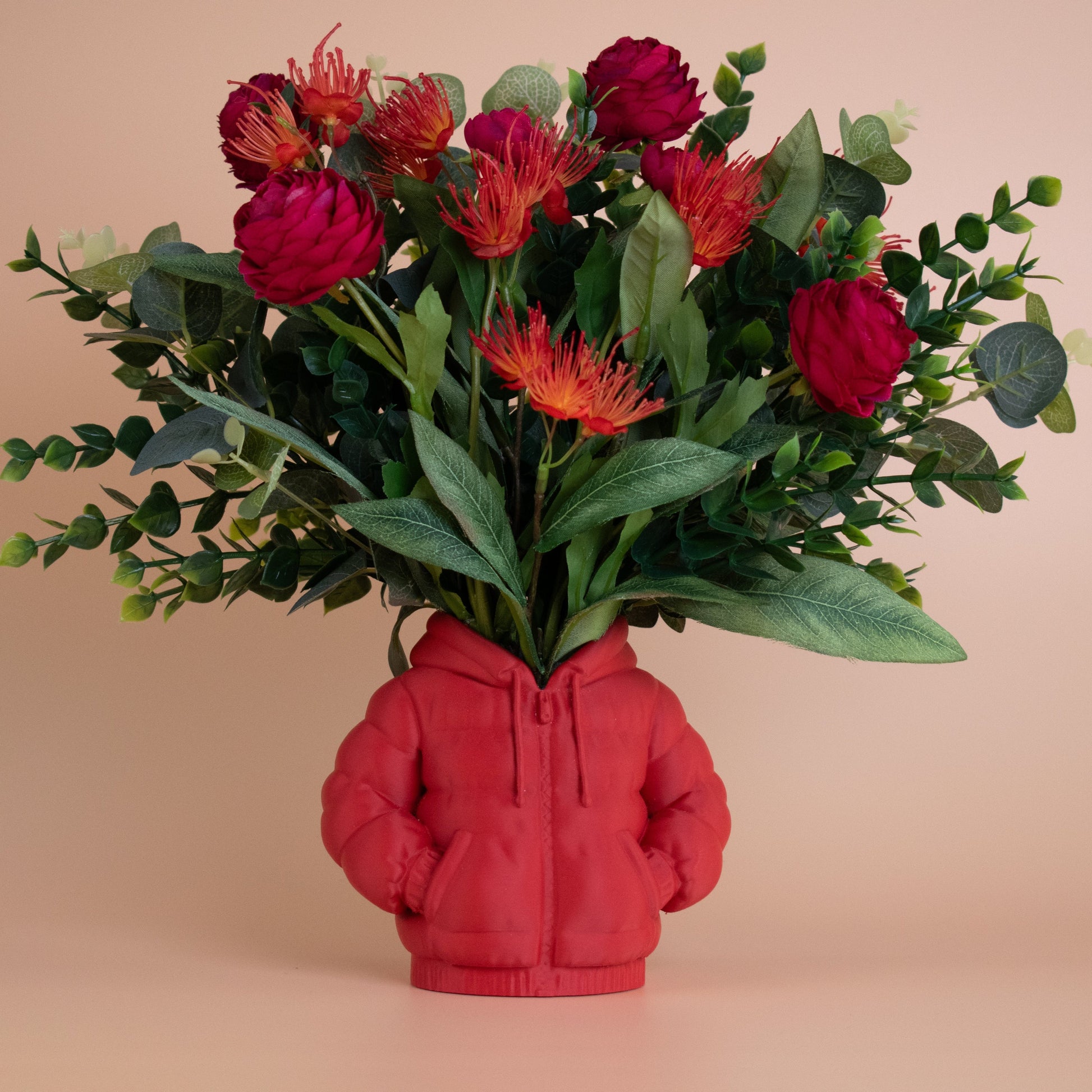 Artificial red and orange floral arrangement in a red hoodie-shaped vase, styled against a warm beige background — a playful, modern décor piece blending fashion and floral design.