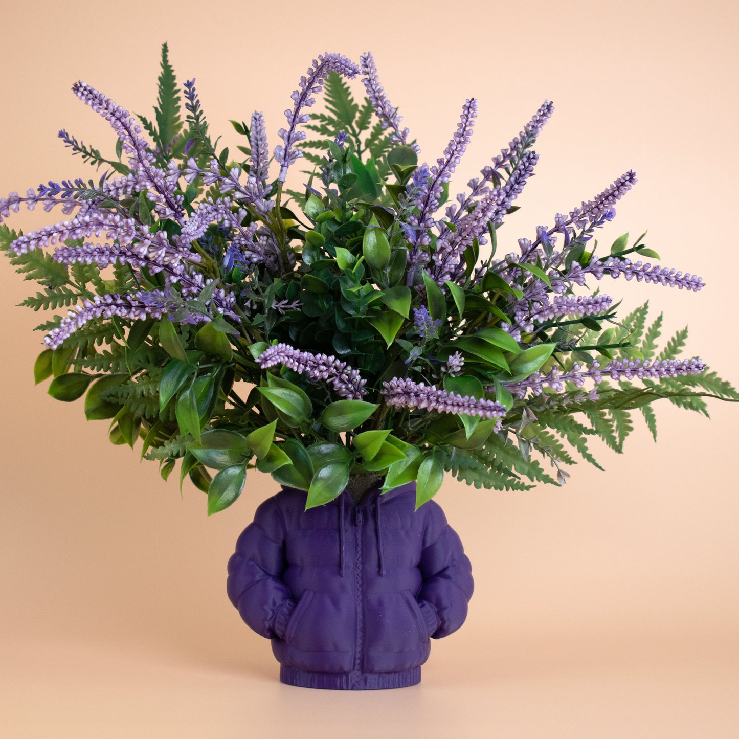 Artificial lavender and green floral arrangement in a purple hoodie-shaped vase against a beige background — a stylish fusion of fashion and modern décor.