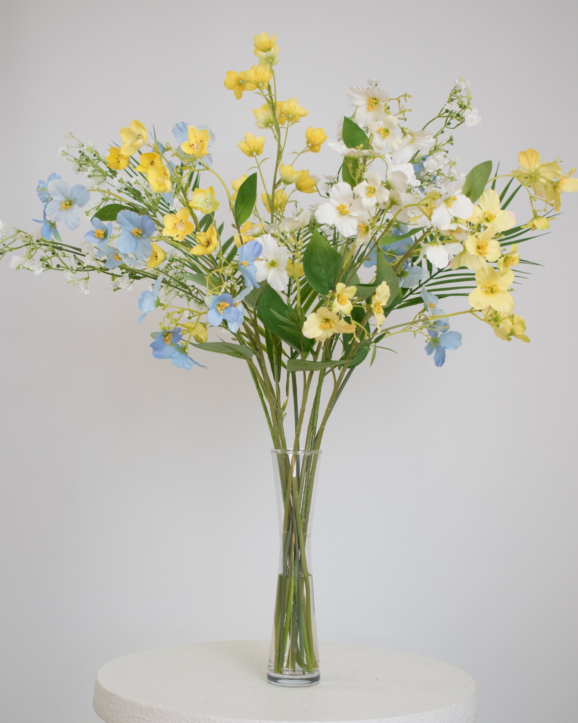 Artificial pastel floral arrangement with yellow, blue, and white flowers, arranged in a tall clear glass vase.