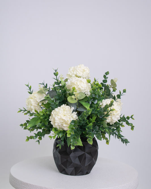 Artificial floral arrangement featuring white hydrangeas and green foliage in a matte black geometric vase, displayed on a white pedestal against a neutral background.