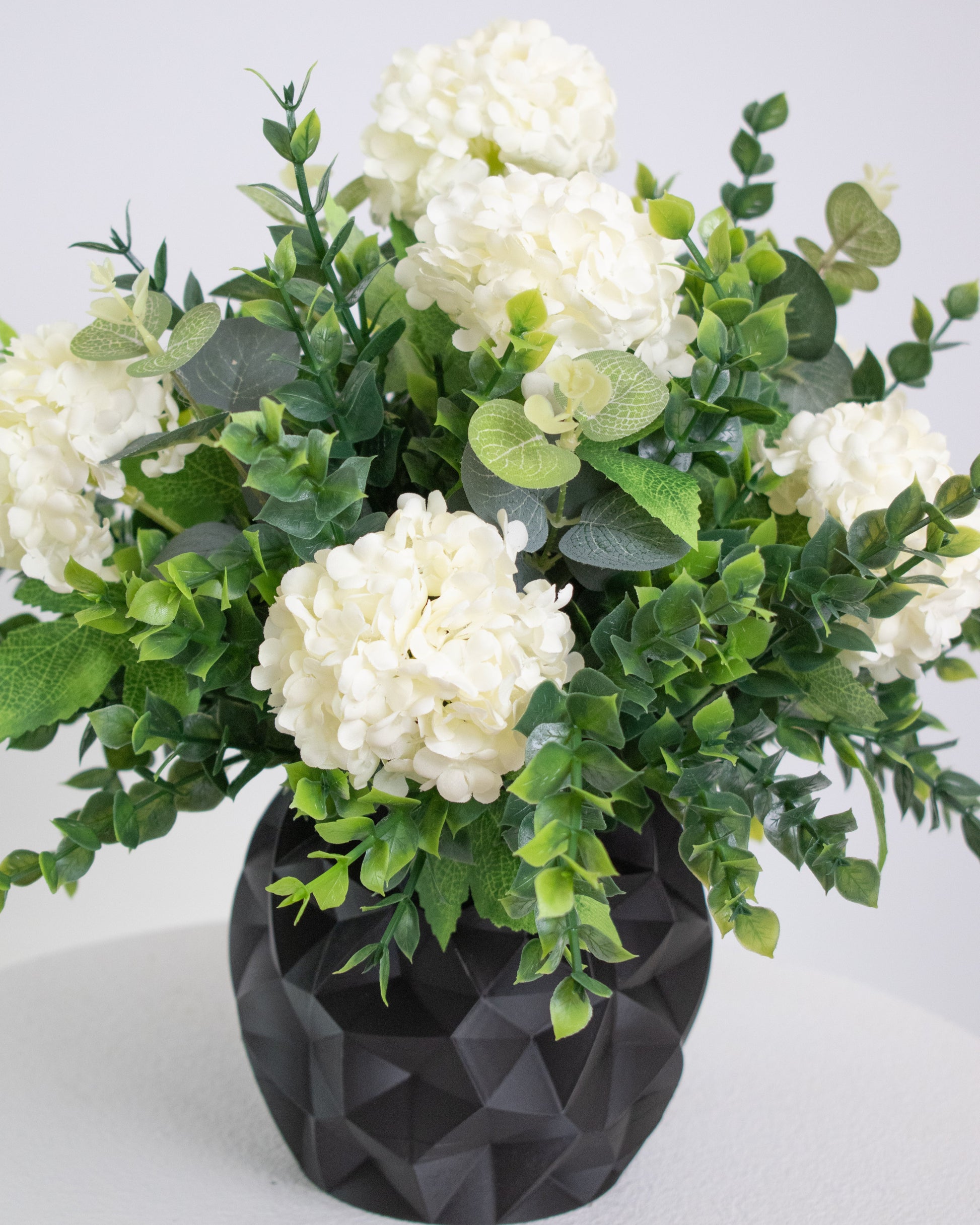 Artificial floral arrangement featuring white hydrangeas and green foliage in a matte black geometric vase, displayed on a white pedestal against a neutral background.