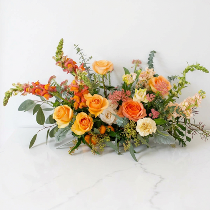 A vibrant floral arrangement featuring orange and yellow roses, peach snapdragons, white chrysanthemums, and green foliage on a white surface against a white background.