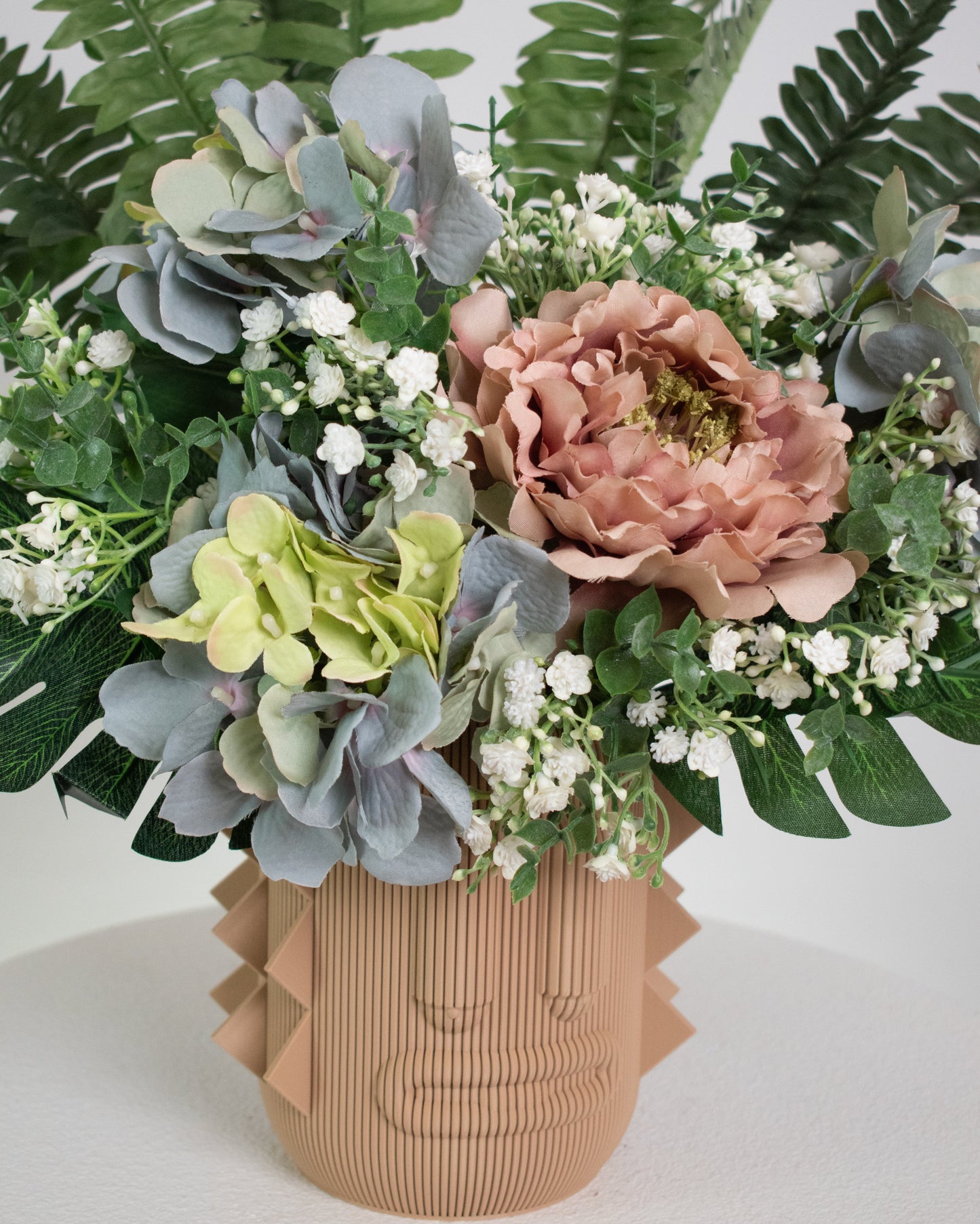 Artificial arrangement with lush ferns, blush peonies, hydrangeas, and white baby’s breath in a terracotta sculptural vase, styled on a marble pedestal against a light background.