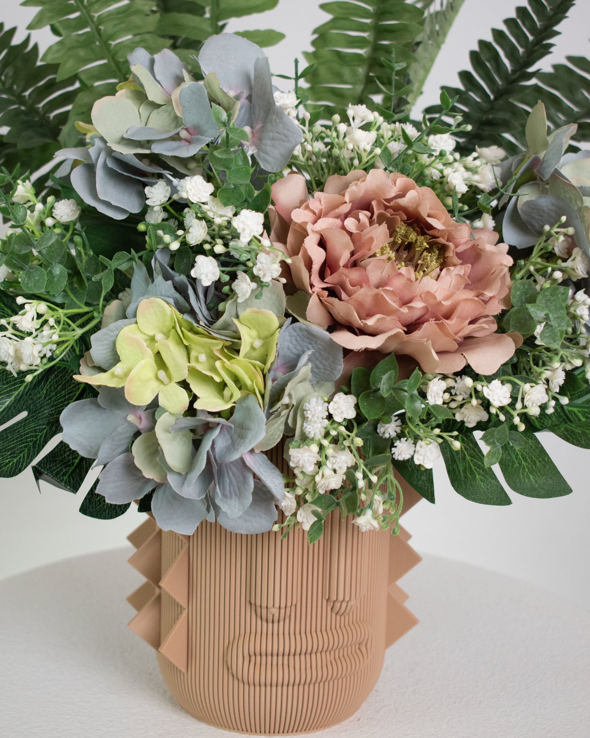 Artificial arrangement with lush ferns, blush peonies, hydrangeas, and white baby’s breath in a terracotta sculptural vase, styled on a marble pedestal against a light background.