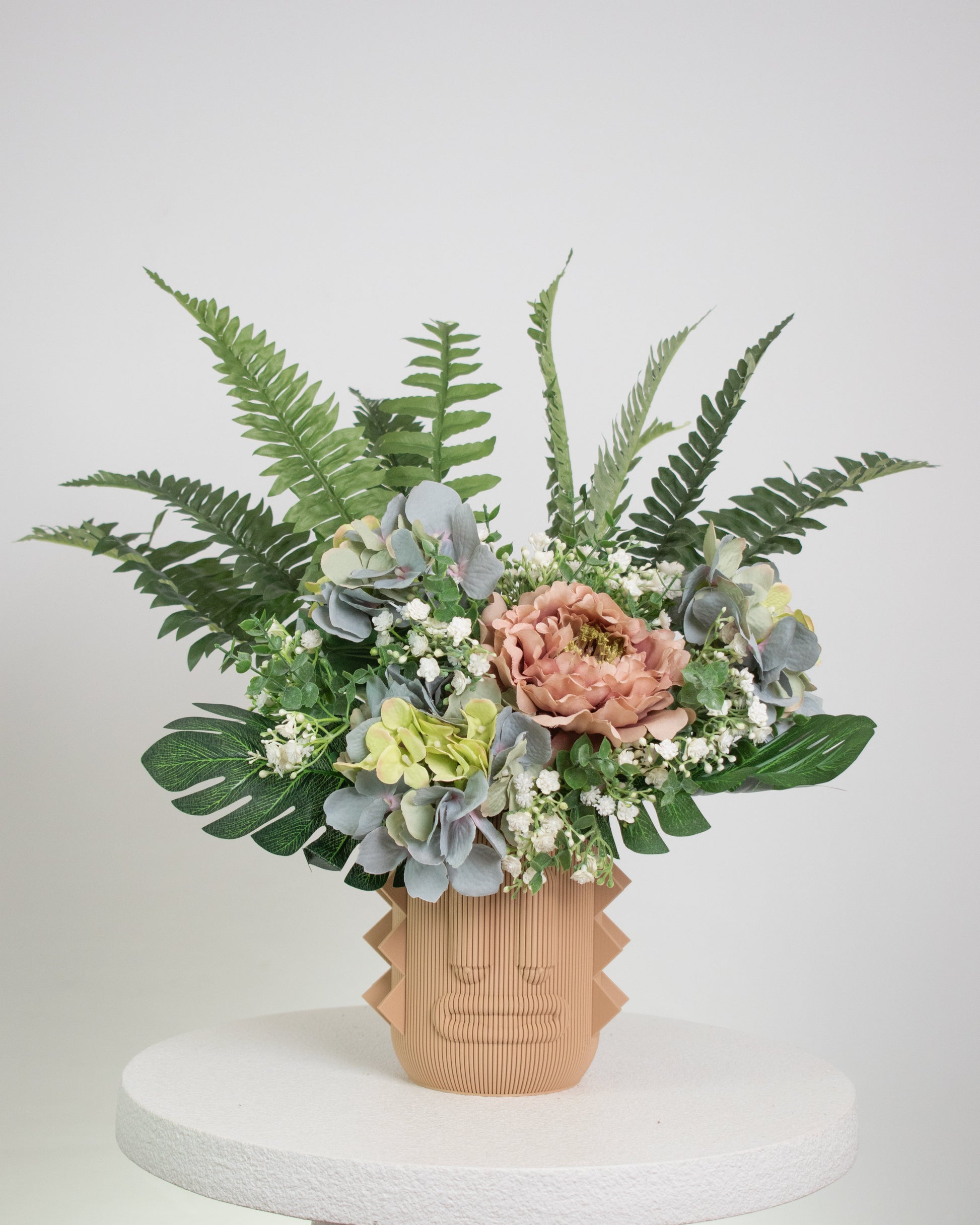 Artificial arrangement with lush ferns, blush peonies, hydrangeas, and white baby’s breath in a terracotta sculptural vase, styled on a marble pedestal against a light background.