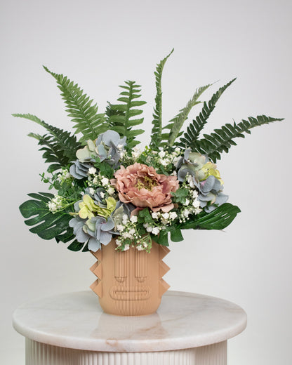 Artificial arrangement with lush ferns, blush peonies, hydrangeas, and white baby’s breath in a terracotta sculptural vase, styled on a marble pedestal against a light background.