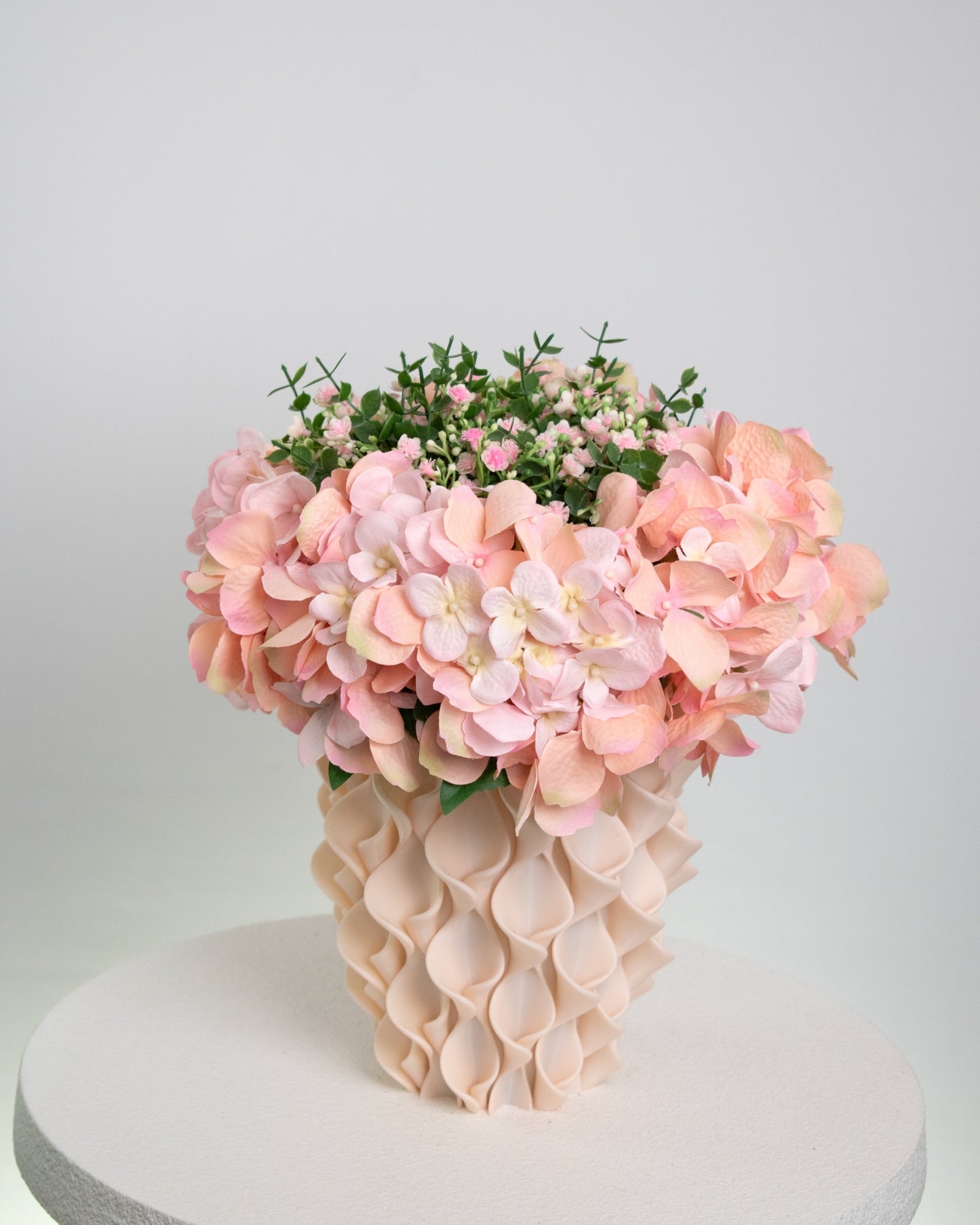 Artificial pink hydrangea and rose arrangement with lush greenery in a sculptural cream vase, styled on a white round pedestal against a soft neutral background.