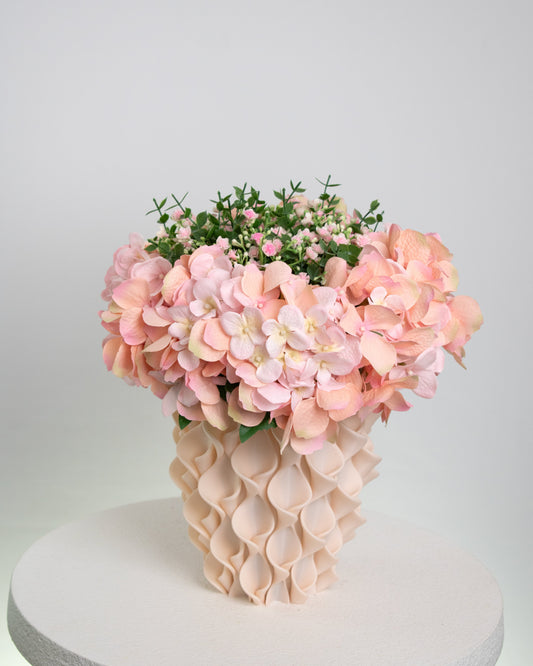 Artificial pink hydrangea and rose arrangement with lush greenery in a sculptural cream vase, styled on a white round pedestal against a soft neutral background.