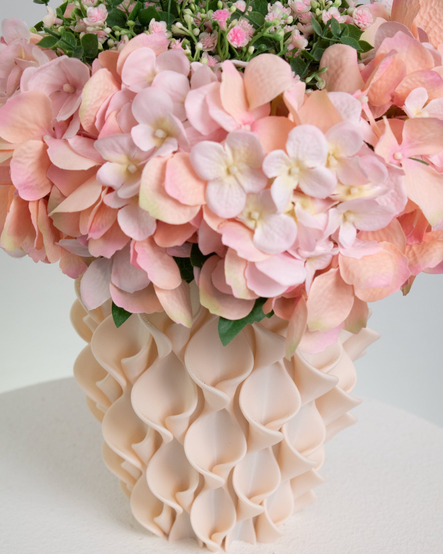 Artificial pink hydrangea and rose arrangement with lush greenery in a sculptural cream vase, styled on a white round pedestal against a soft neutral background.