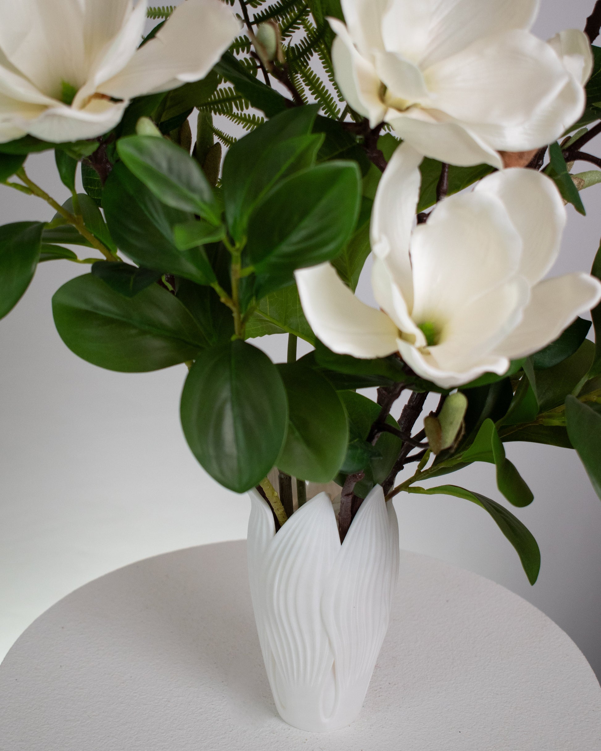 Artificial white magnolia flower arrangement with lush green leaves and fern accents, beautifully styled in a sleek white vase on a round white table, set against a neutral background.