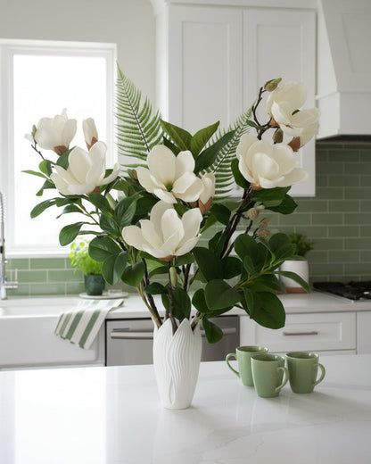 Artificial white magnolia flower arrangement with lush green leaves and fern accents, beautifully styled in a sleek white vase on a round white table, set against a neutral background.