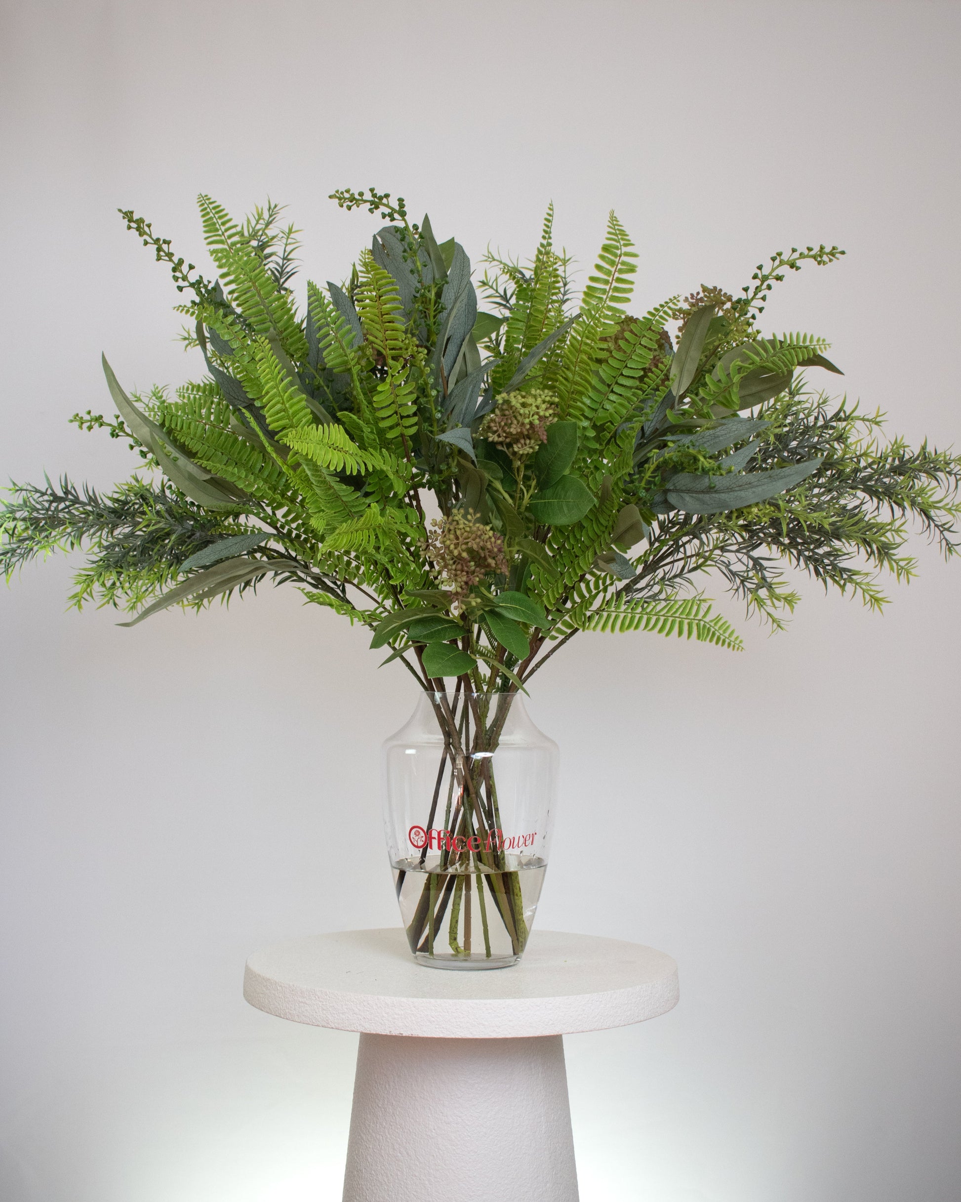 Artificial green foliage arrangement with ferns and leafy stems, displayed in a clear glass vase with acrylic water
