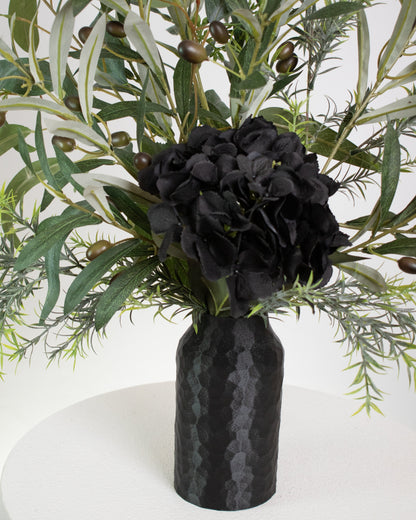 Artificial floral arrangement featuring lifelike olive branches with dark berries and a black hydrangea bloom, elegantly displayed in a textured black vase on a marble-topped round table against a neutral background.