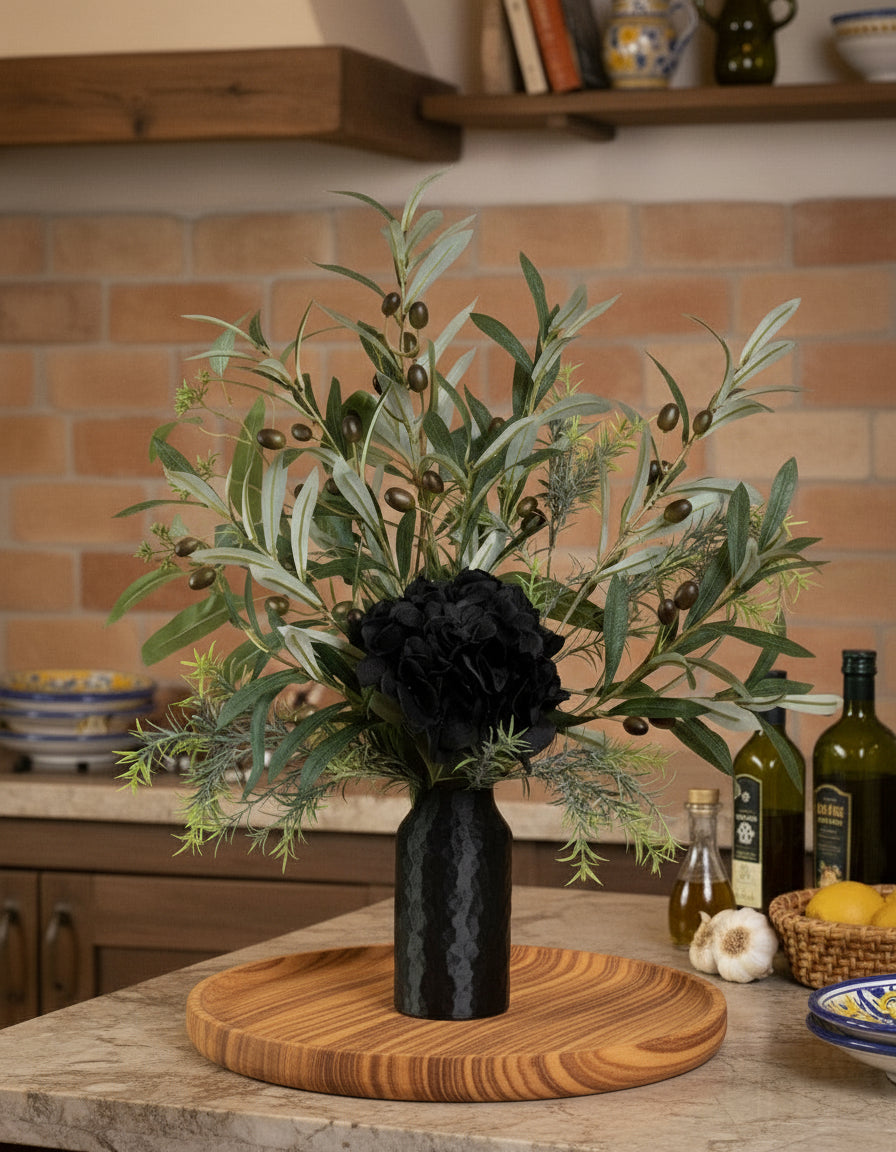 Artificial floral arrangement featuring lifelike olive branches with dark berries and a black hydrangea bloom, elegantly displayed in a textured black vase on a marble-topped round table against a neutral background.