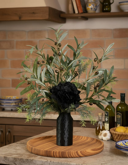 Artificial floral arrangement featuring lifelike olive branches with dark berries and a black hydrangea bloom, elegantly displayed in a textured black vase on a marble-topped round table against a neutral background.