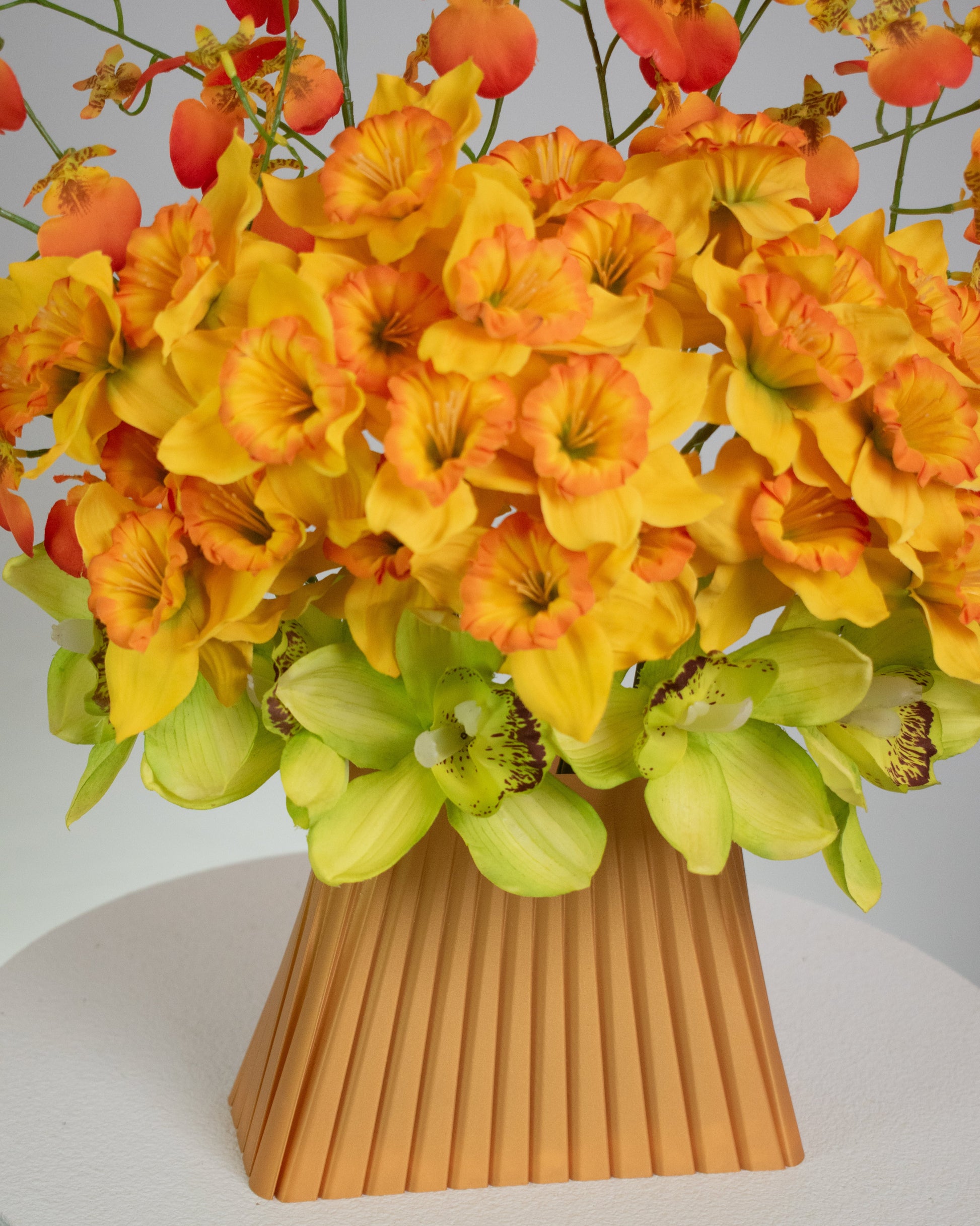 Vibrant artificial floral arrangement featuring orange orchids, yellow blossoms, and green cymbidiums in a pleated terracotta vase, styled on a white pedestal against a soft neutral background.