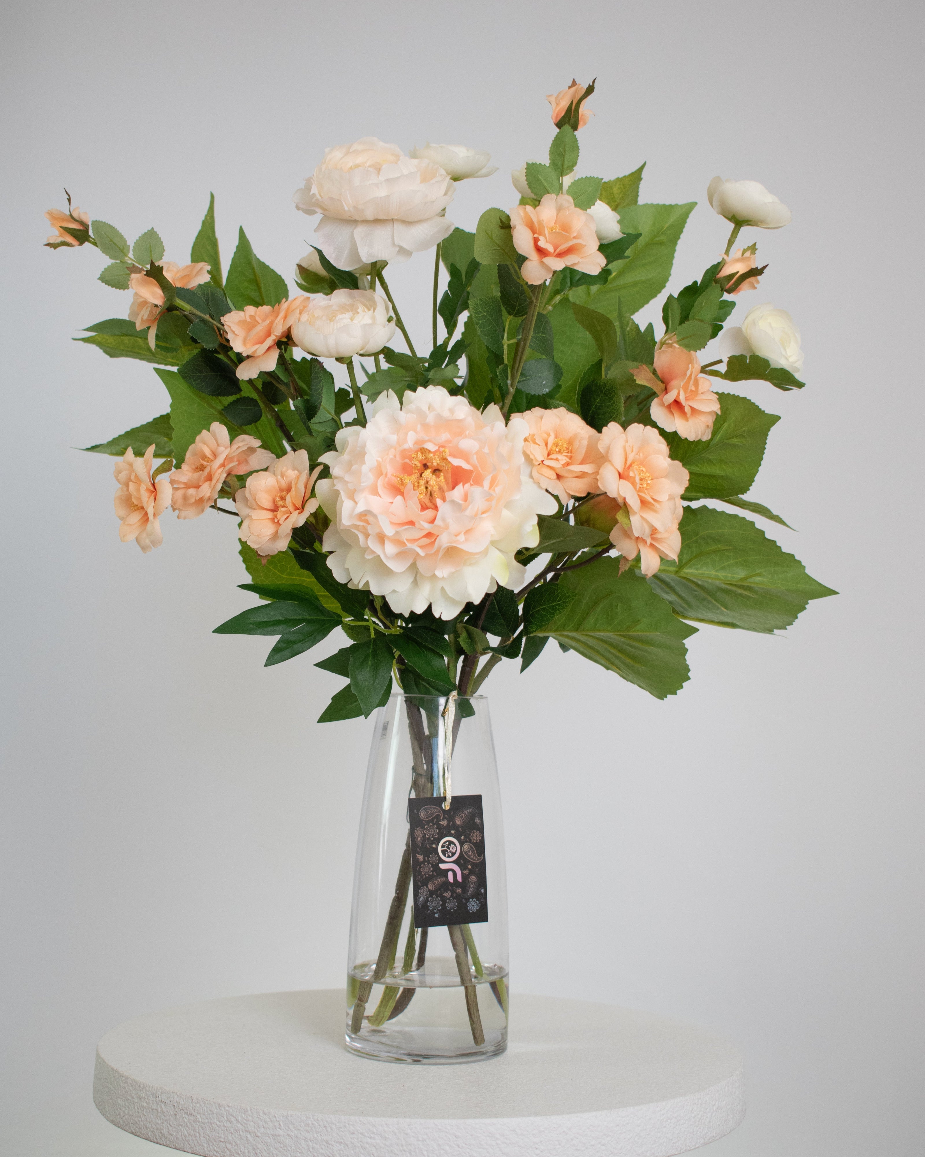 Bouquet of peach and white flowers in a clear vase on a white pedestal against a light gray background