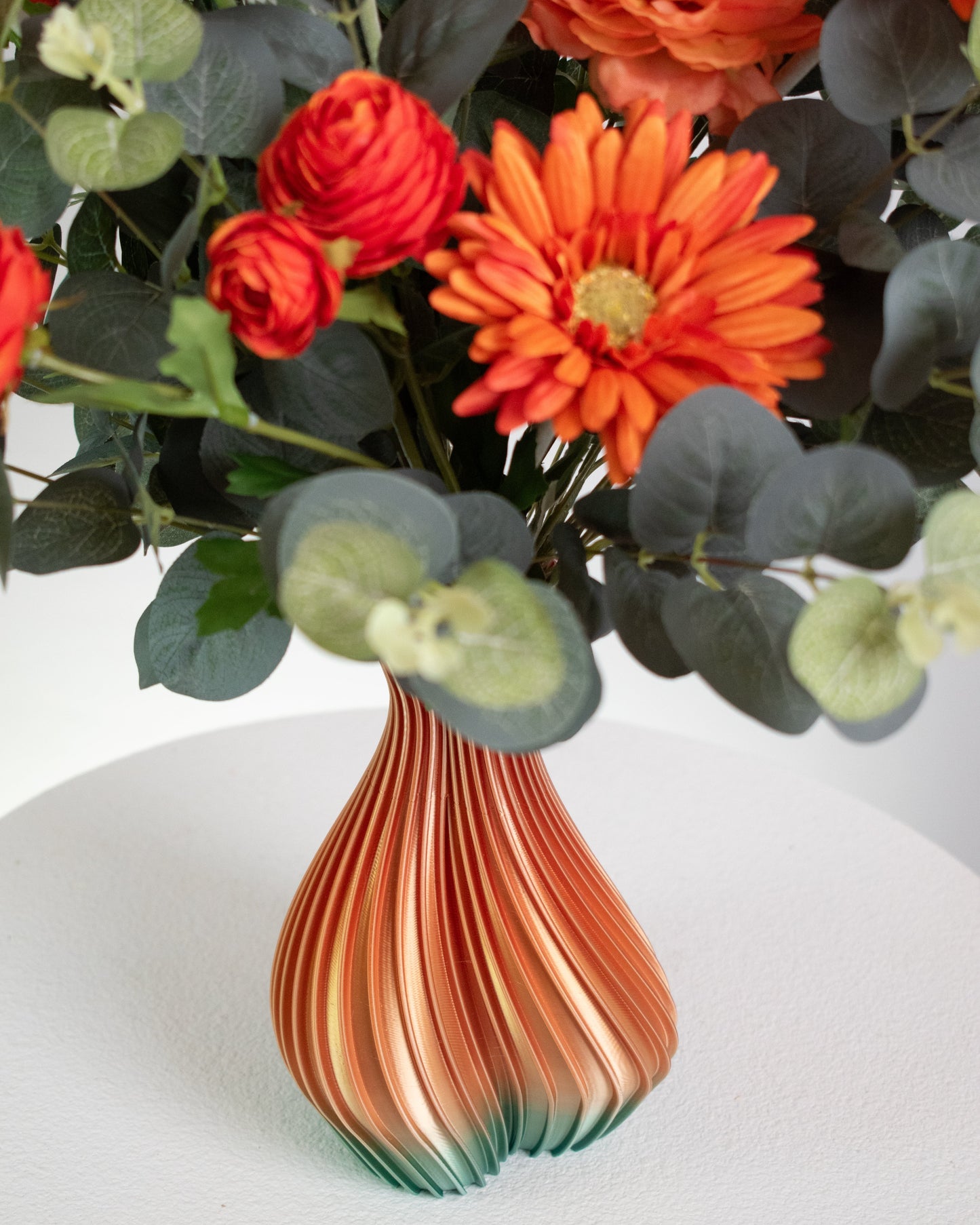 Artificial floral arrangement with orange gerberas and ranunculus surrounded by eucalyptus leaves, displayed in an ombre vase blending amber and green tones on a light background.