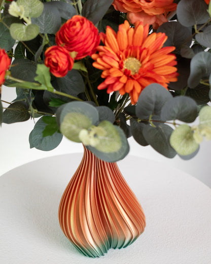 Artificial floral arrangement with orange gerberas and ranunculus surrounded by eucalyptus leaves, displayed in an ombre vase blending amber and green tones on a light background.