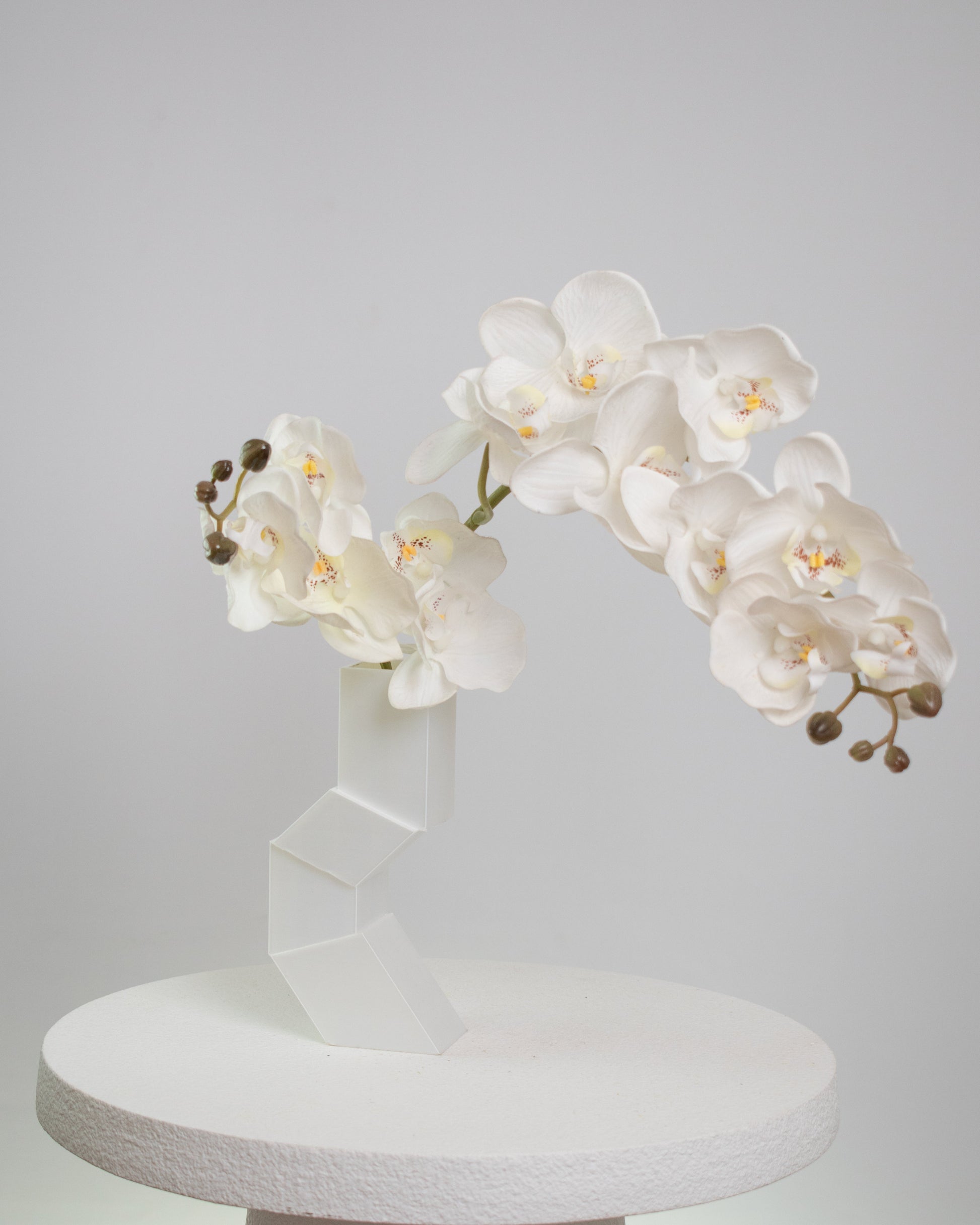 Artificial white orchid arrangement cascading gracefully from a modern geometric white vase, displayed on a white pedestal against a minimalist neutral background.
