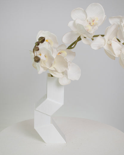 Artificial white orchid arrangement cascading gracefully from a modern geometric white vase, displayed on a white pedestal against a minimalist neutral background.