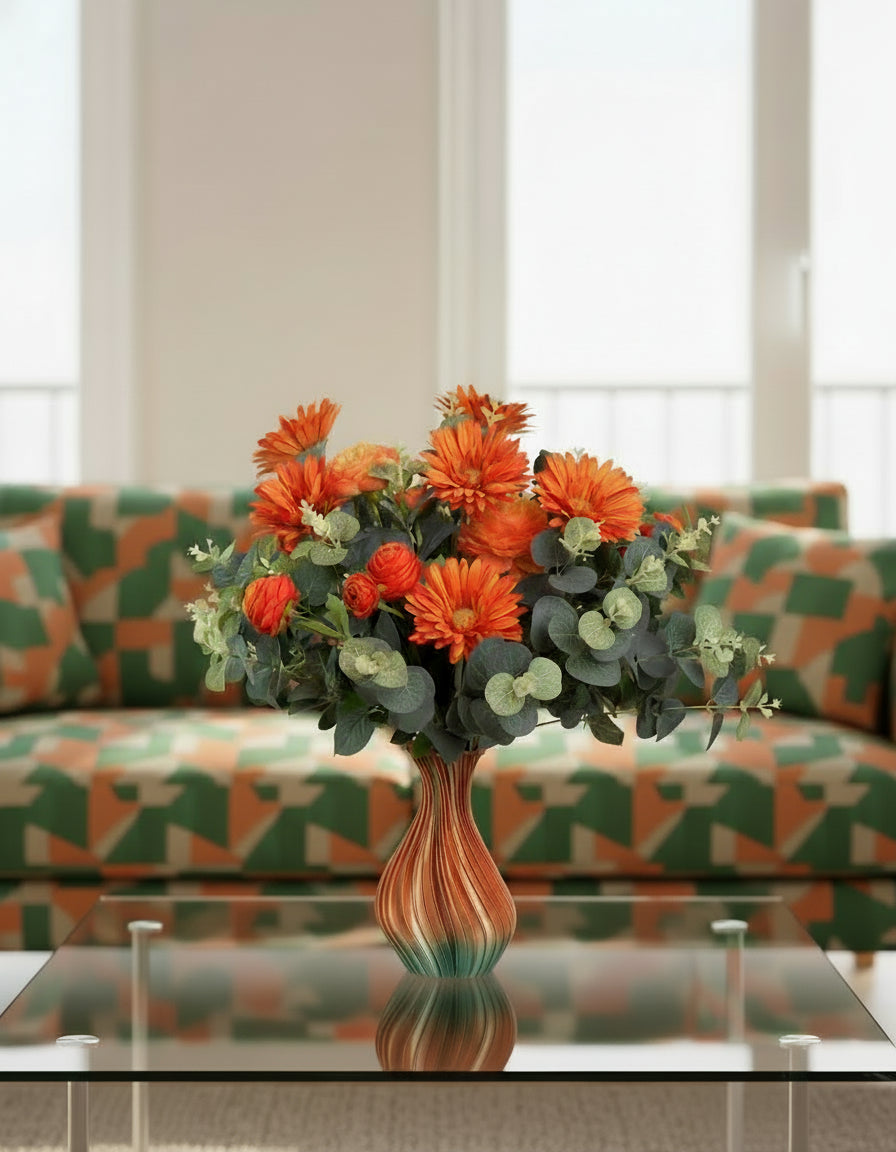 Artificial floral arrangement with orange gerberas and ranunculus surrounded by eucalyptus leaves, displayed in an ombre vase blending amber and green tones on a light background.