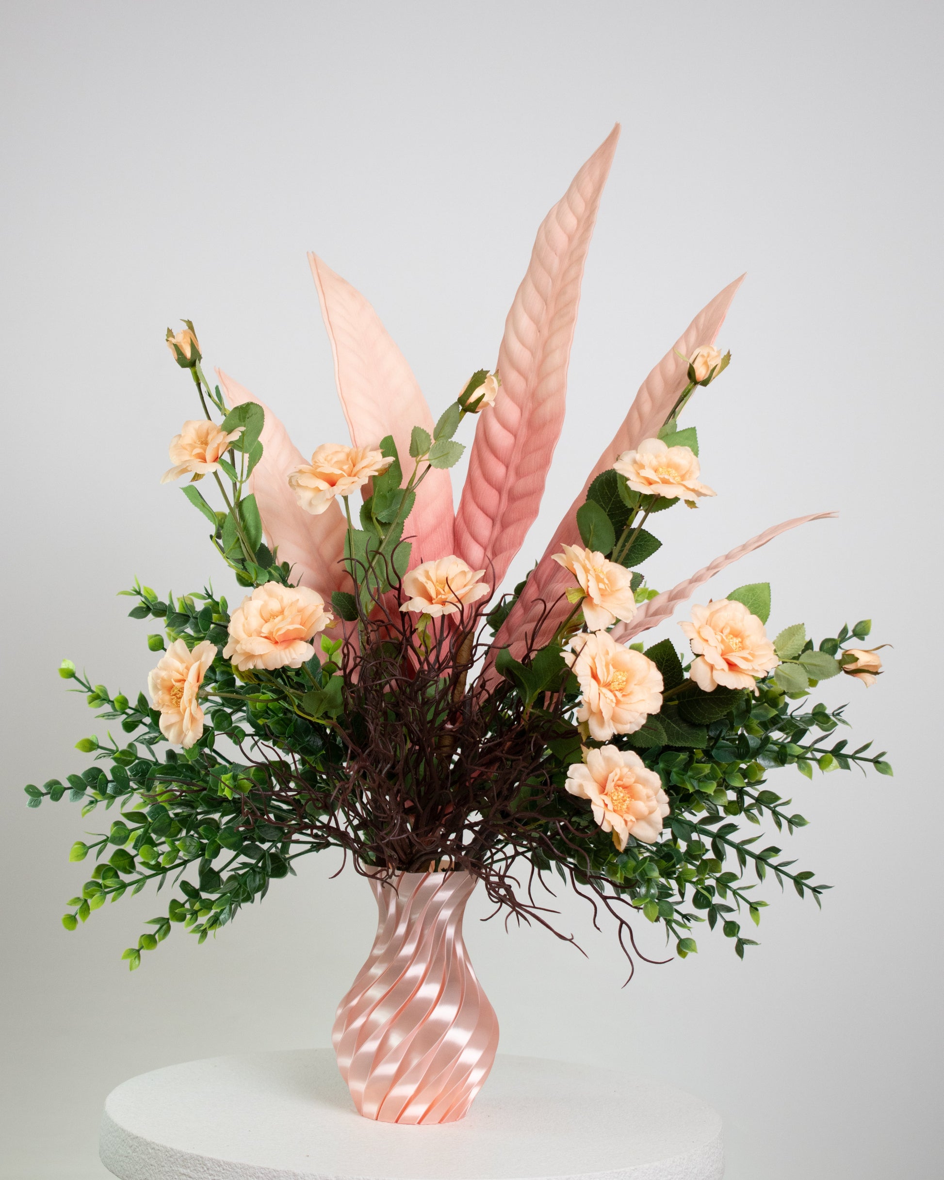 Artificial floral arrangement with peach roses, pink tropical leaves, and lush greenery in a twisted pink spiral vase, displayed on a white pedestal — perfect for elegant or modern interior spaces.