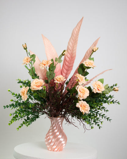 Artificial floral arrangement with peach roses, pink tropical leaves, and lush greenery in a twisted pink spiral vase, displayed on a white pedestal — perfect for elegant or modern interior spaces.