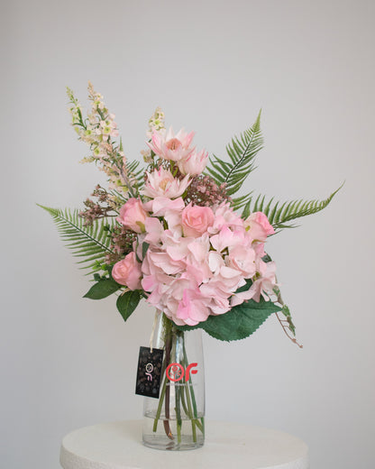Artificial pink floral arrangement with hydrangeas, roses, and ferns, arranged in a clear glass vase with acrylic water.