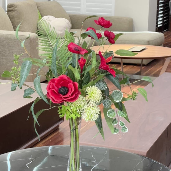 Artificial red floral arrangement with lush greenery and cream accents, displayed in a tall clear glass vase with acrylic water