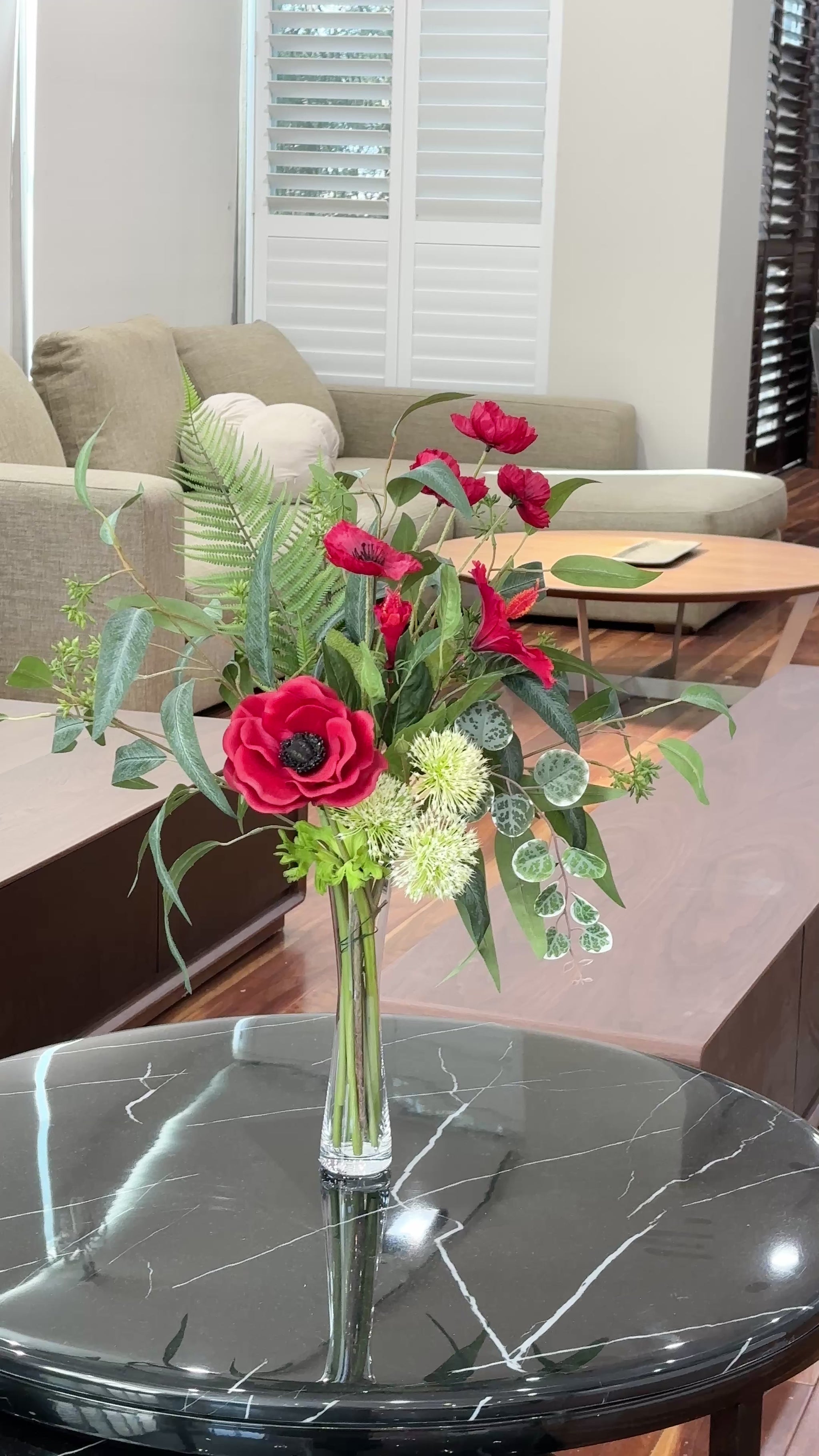 Artificial red floral arrangement with lush greenery and cream accents, displayed in a tall clear glass vase with acrylic water