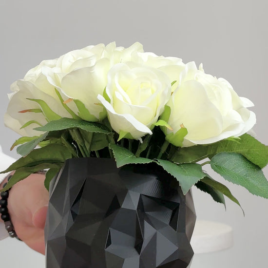 Artificial ivory rose arrangement displayed in a black geometric skull vase on a white pedestal against a neutral background.