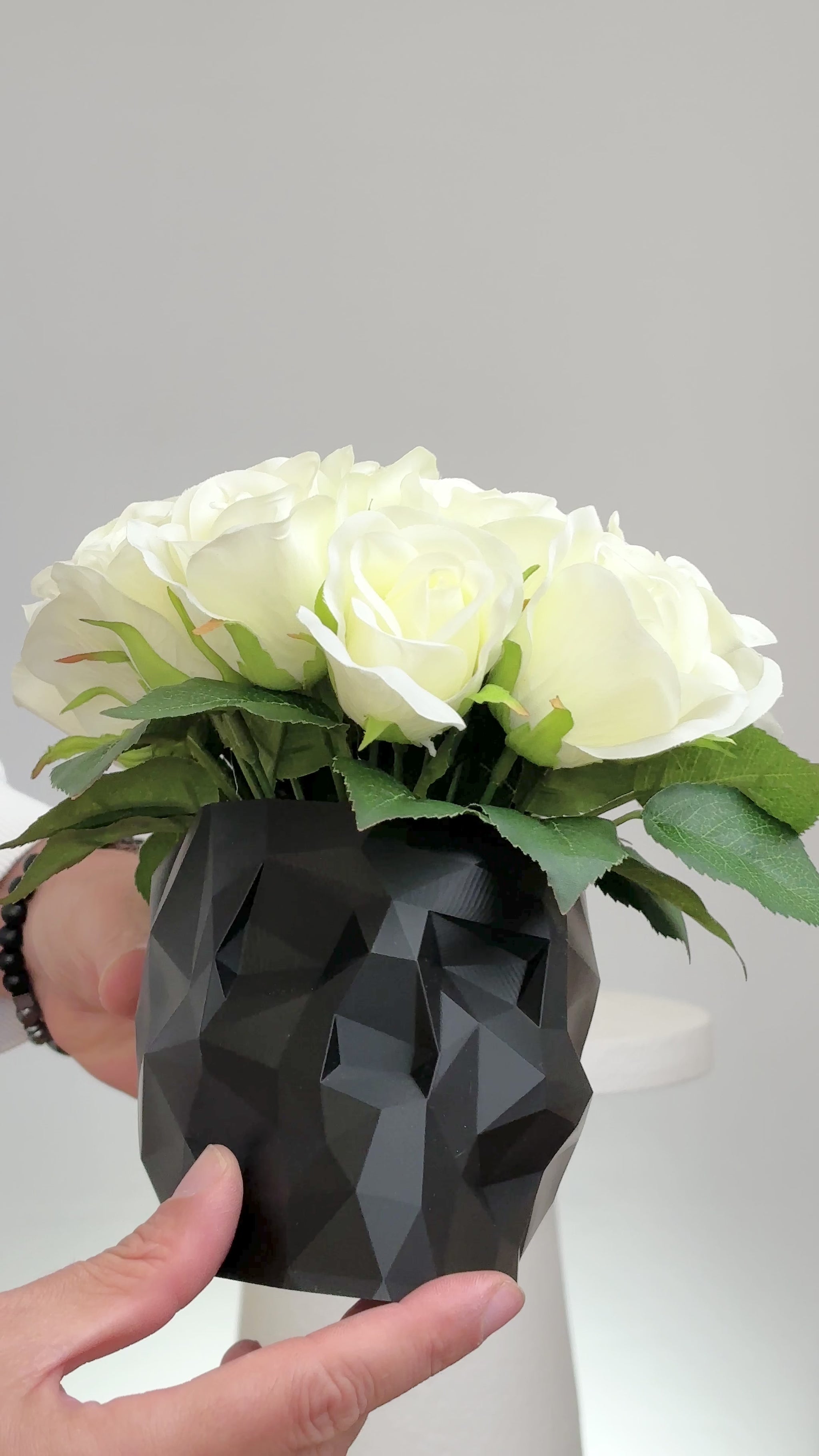 Artificial ivory rose arrangement displayed in a black geometric skull vase on a white pedestal against a neutral background.