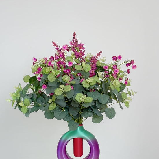 Artificial floral arrangement with pink and purple blossoms and eucalyptus greenery, displayed in a round ombre vase blending purple, green, and rose tones, on a neutral background.