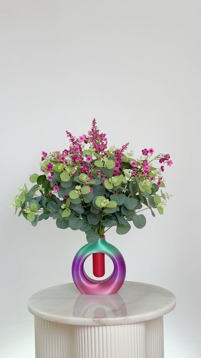 Artificial floral arrangement with pink and purple blossoms and eucalyptus greenery, displayed in a round ombre vase blending purple, green, and rose tones, on a neutral background.