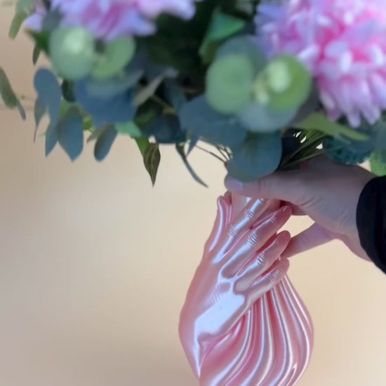 Artificial floral arrangement with pink and white blooms and lush green foliage, elegantly displayed in a sculptural pink hand-shaped vase on a neutral background.