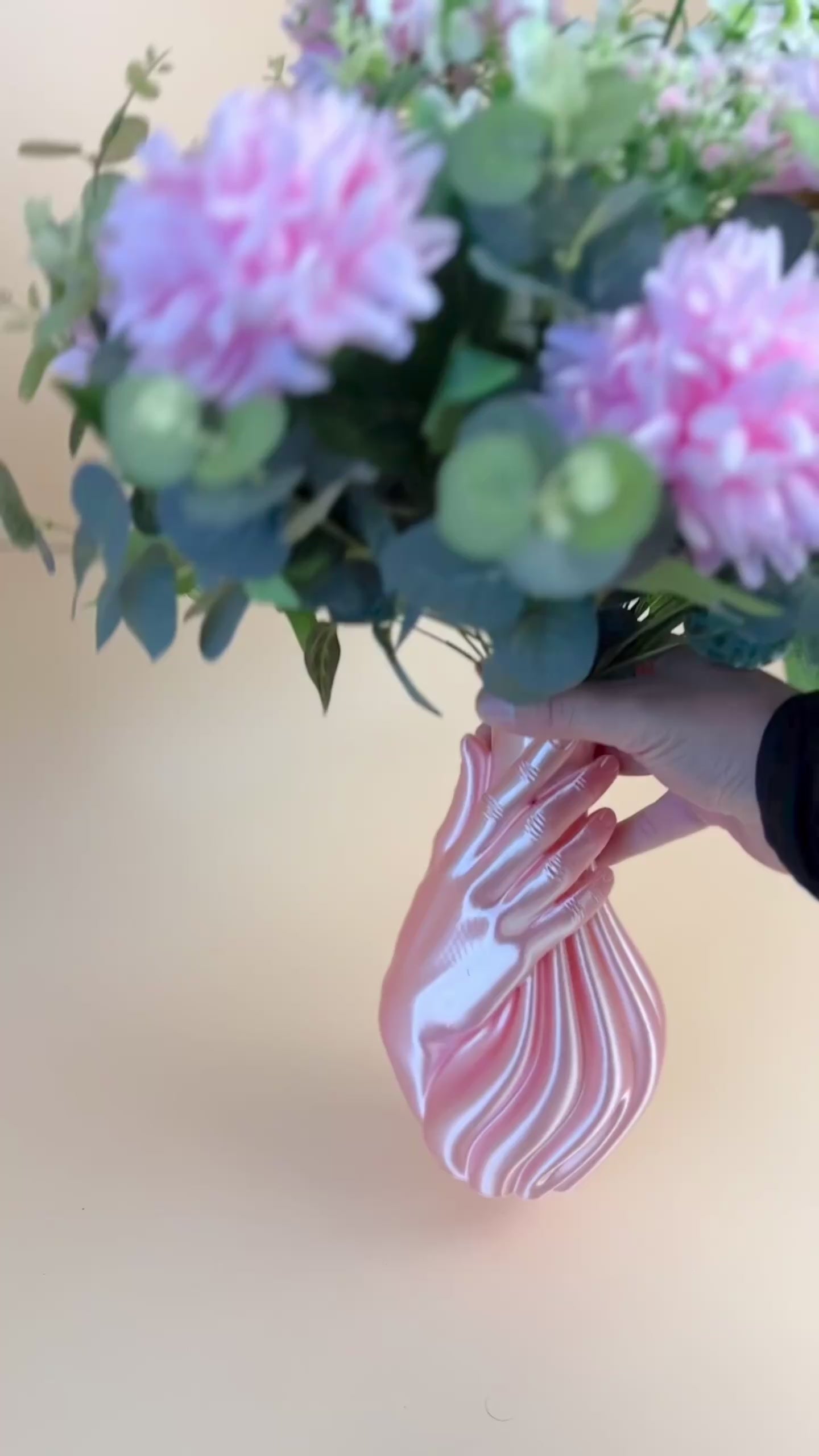 Artificial floral arrangement with pink and white blooms and lush green foliage, elegantly displayed in a sculptural pink hand-shaped vase on a neutral background.