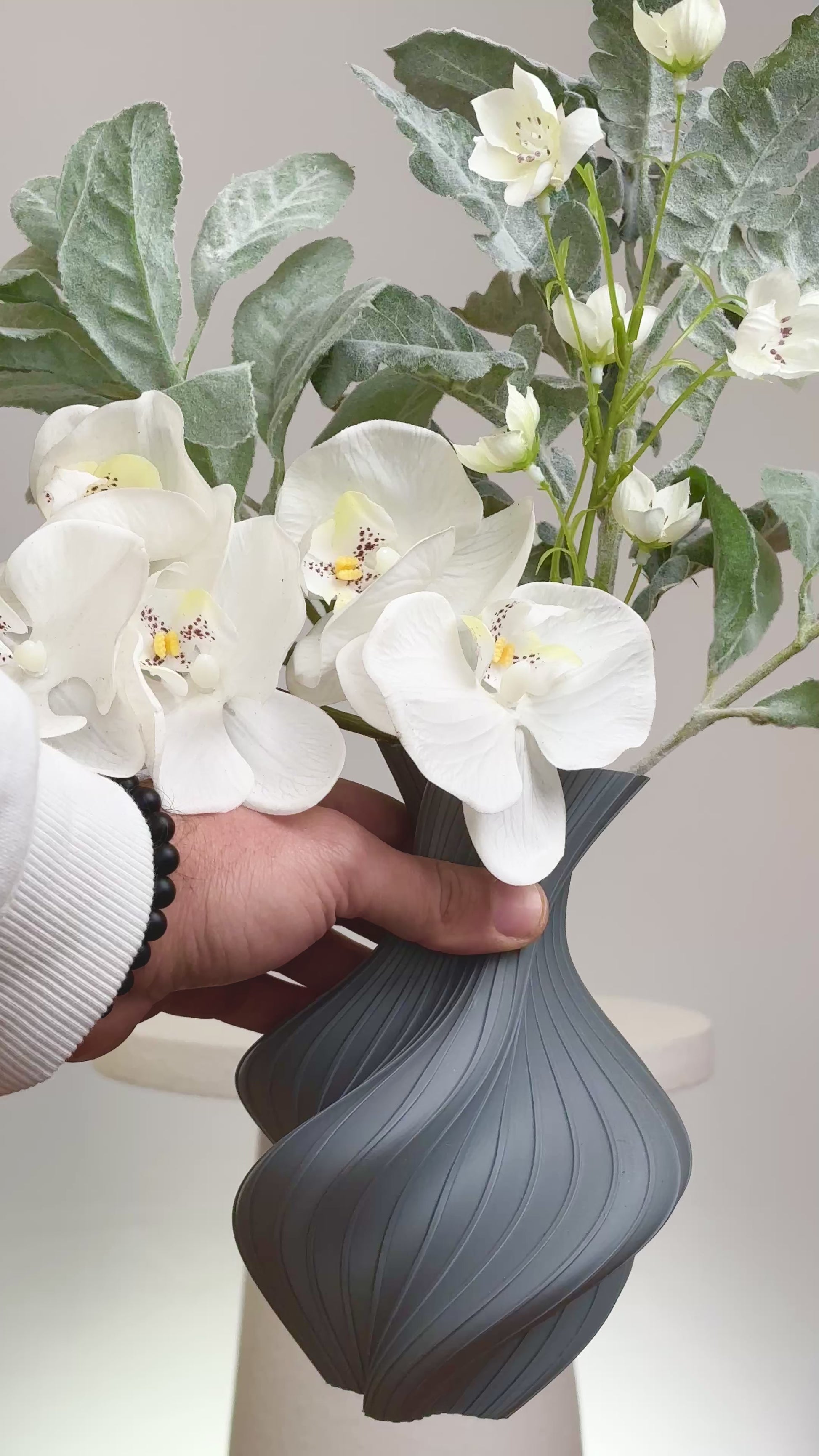 Artificial floral arrangement featuring white orchids and silver-green foliage in a sculptural grey vase with curved lines, displayed on a round white pedestal.