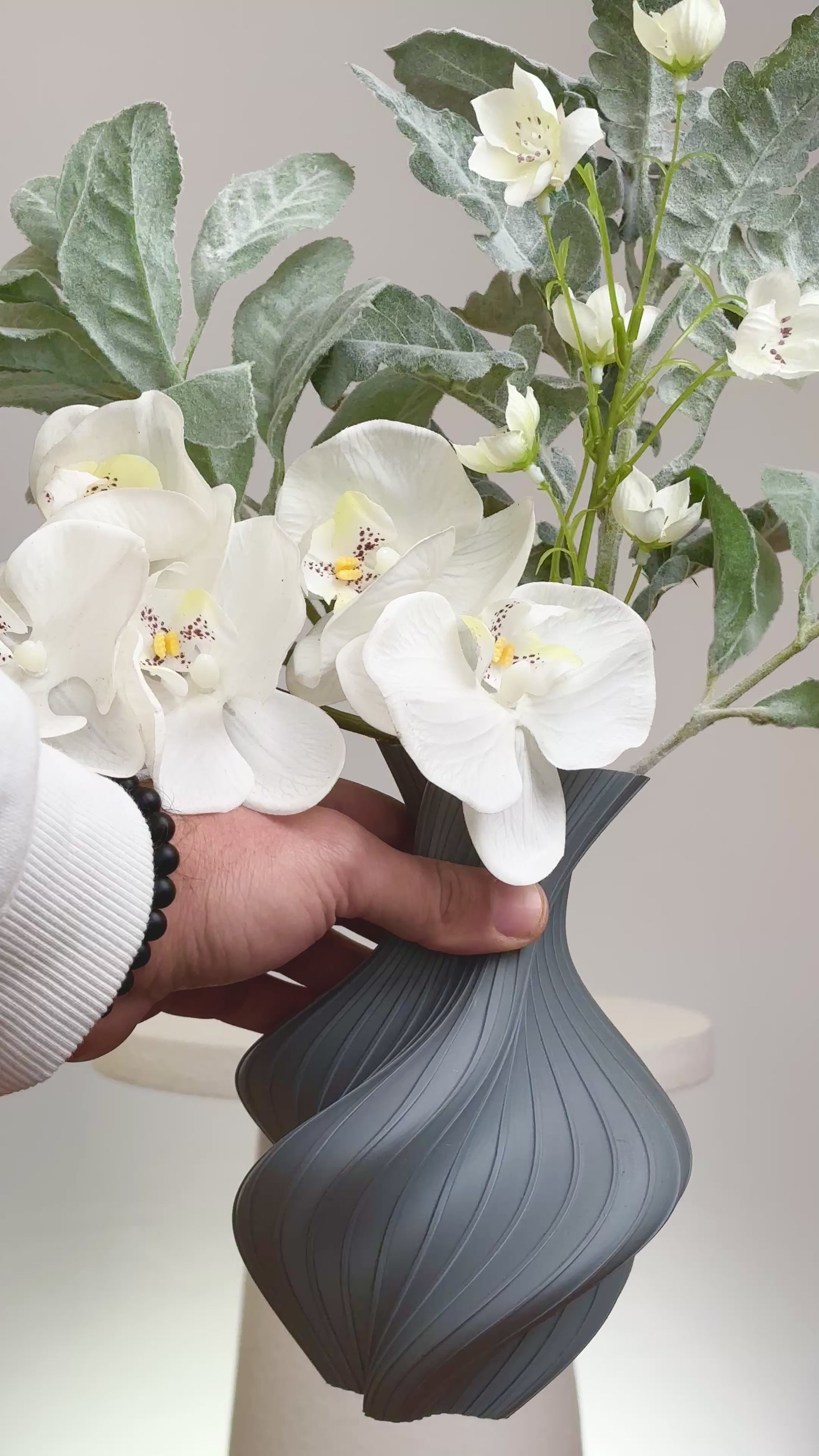 Artificial floral arrangement featuring white orchids and silver-green foliage in a sculptural grey vase with curved lines, displayed on a round white pedestal.