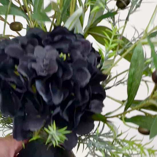 Artificial floral arrangement featuring lifelike olive branches with dark berries and a black hydrangea bloom, elegantly displayed in a textured black vase on a marble-topped round table against a neutral background.