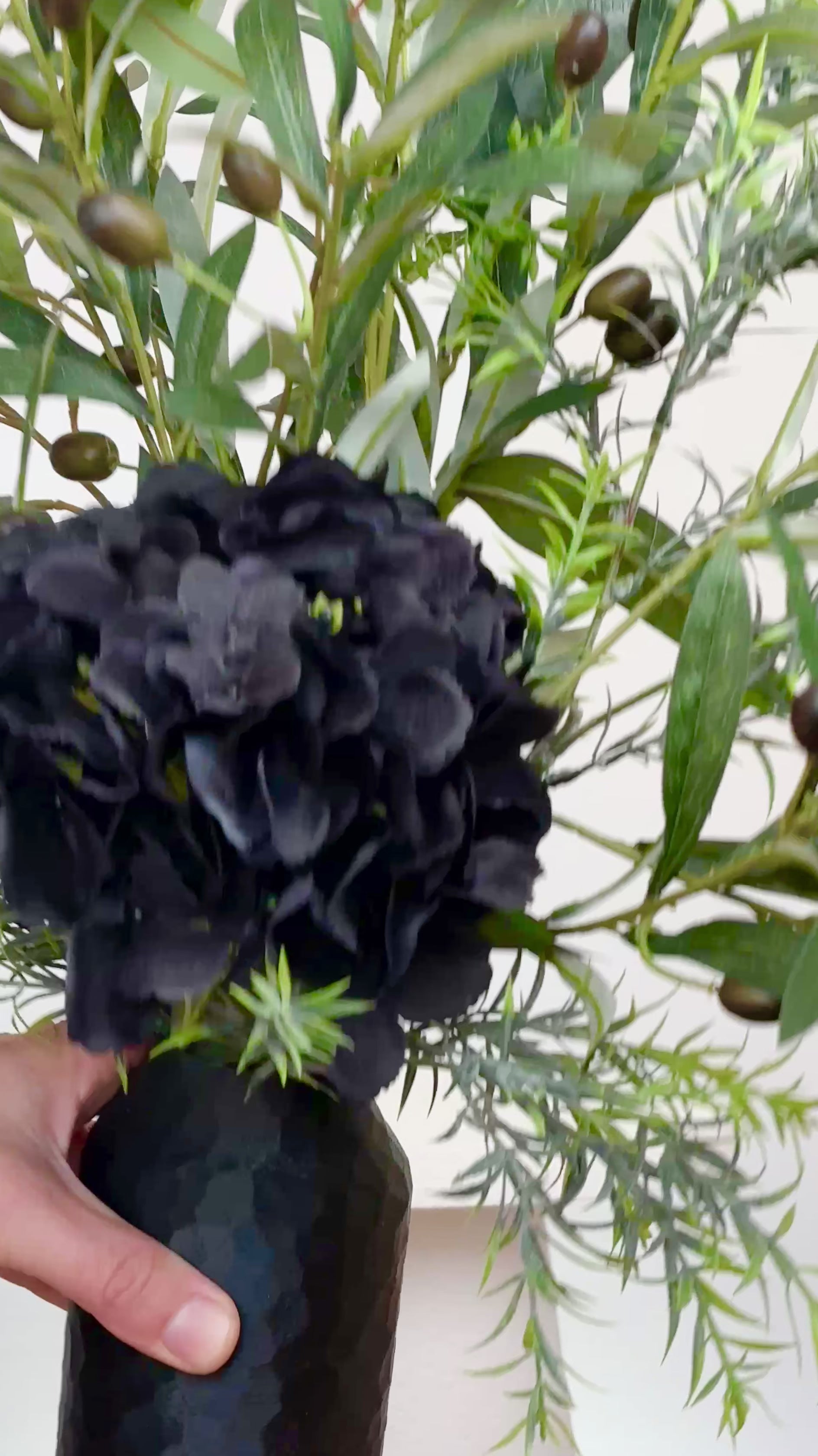 Artificial floral arrangement featuring lifelike olive branches with dark berries and a black hydrangea bloom, elegantly displayed in a textured black vase on a marble-topped round table against a neutral background.
