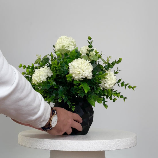 Artificial floral arrangement featuring white hydrangeas and green foliage in a matte black geometric vase, displayed on a white pedestal against a neutral background.