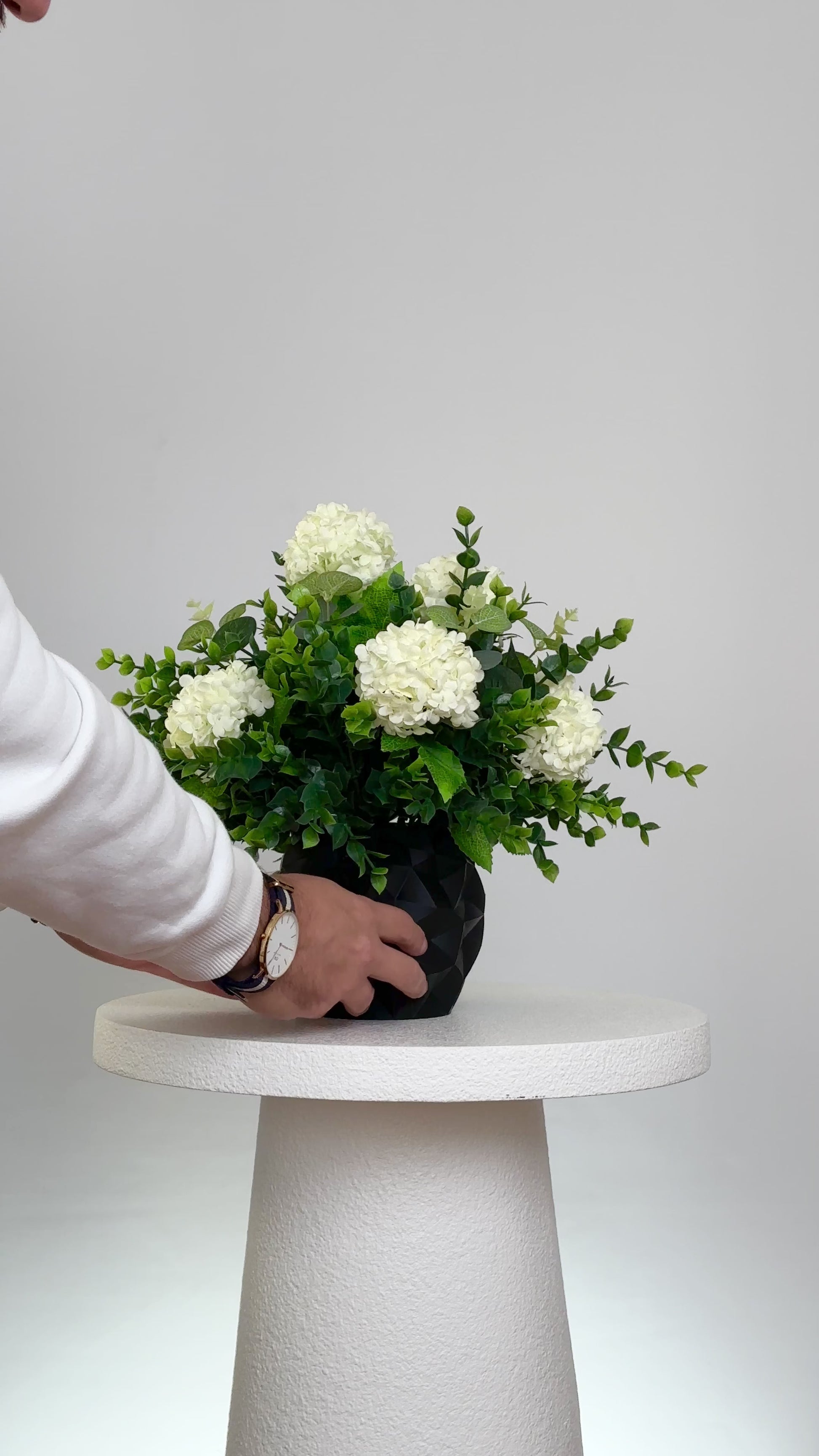 Artificial floral arrangement featuring white hydrangeas and green foliage in a matte black geometric vase, displayed on a white pedestal against a neutral background.