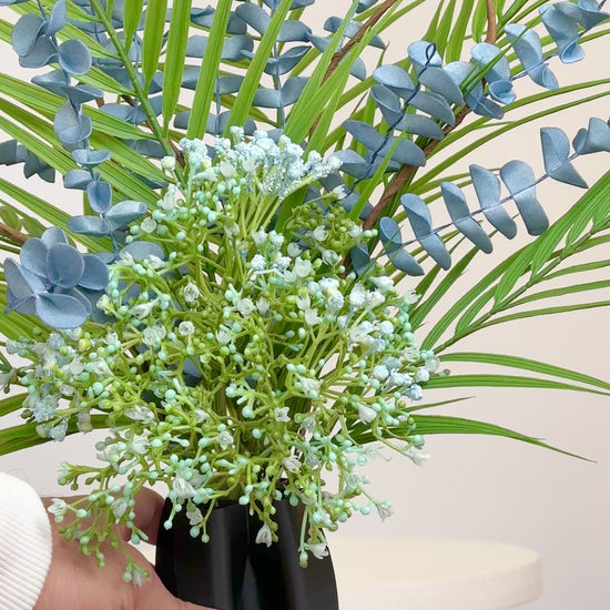Artificial arrangement with blue eucalyptus stems, palm leaves, and delicate greenery in a sleek black sculpted vase, styled on a white pedestal — ideal for modern or minimalist interiors with cool or neutral tones.