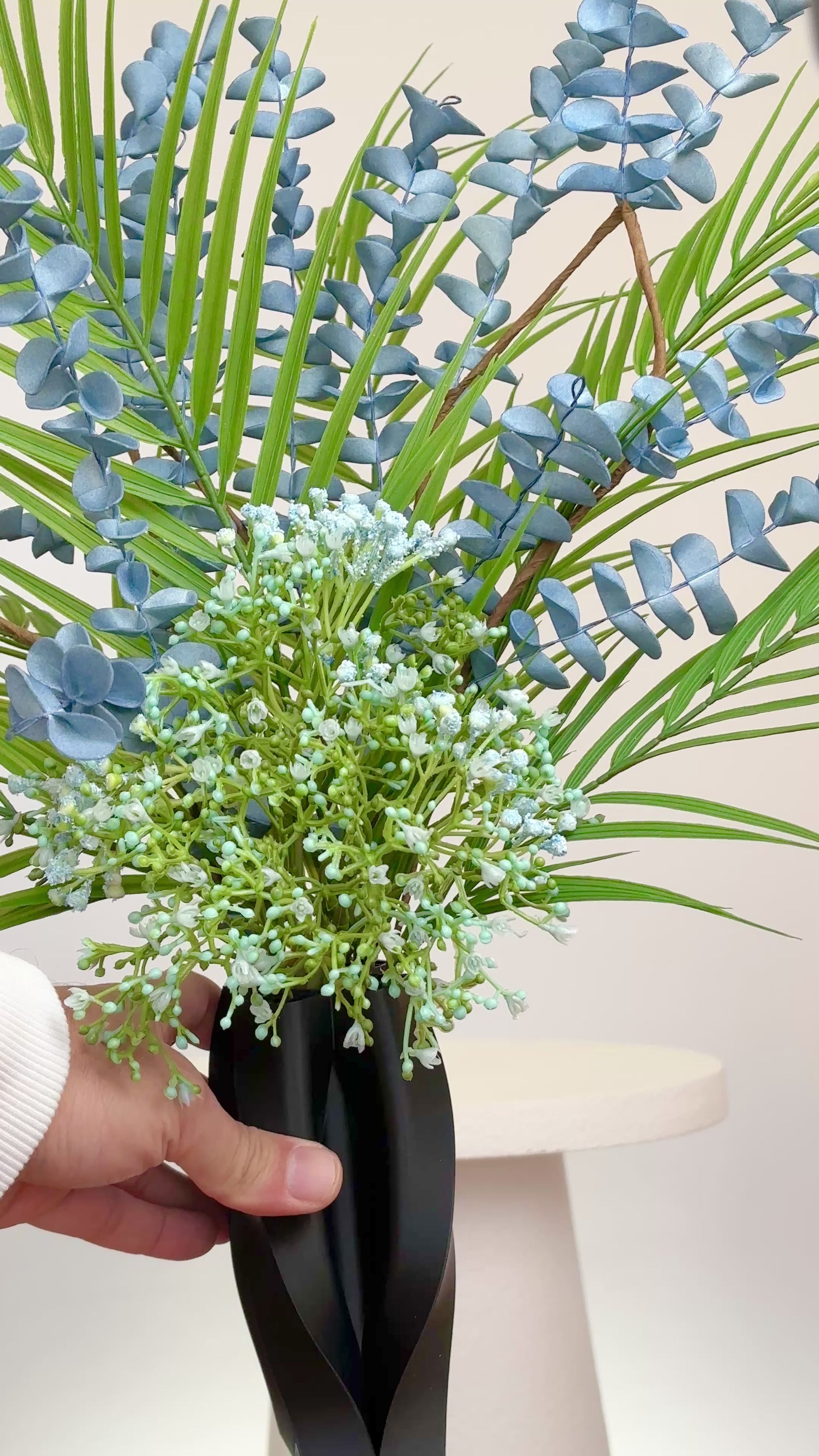 Artificial arrangement with blue eucalyptus stems, palm leaves, and delicate greenery in a sleek black sculpted vase, styled on a white pedestal — ideal for modern or minimalist interiors with cool or neutral tones.