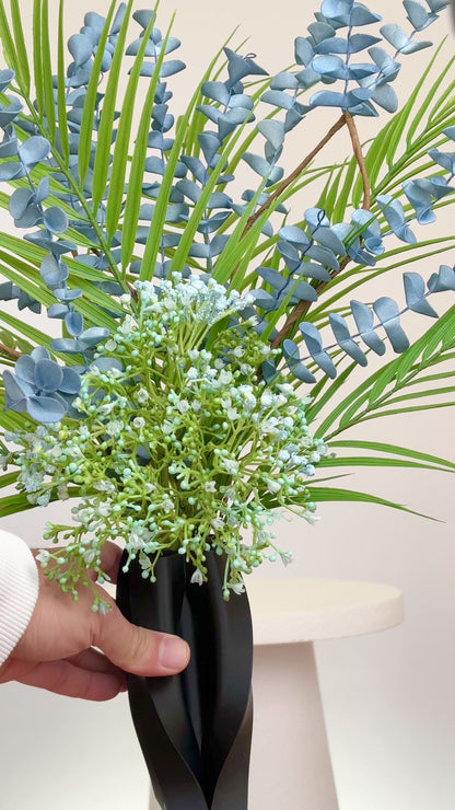 Artificial arrangement with blue eucalyptus stems, palm leaves, and delicate greenery in a sleek black sculpted vase, styled on a white pedestal — ideal for modern or minimalist interiors with cool or neutral tones.