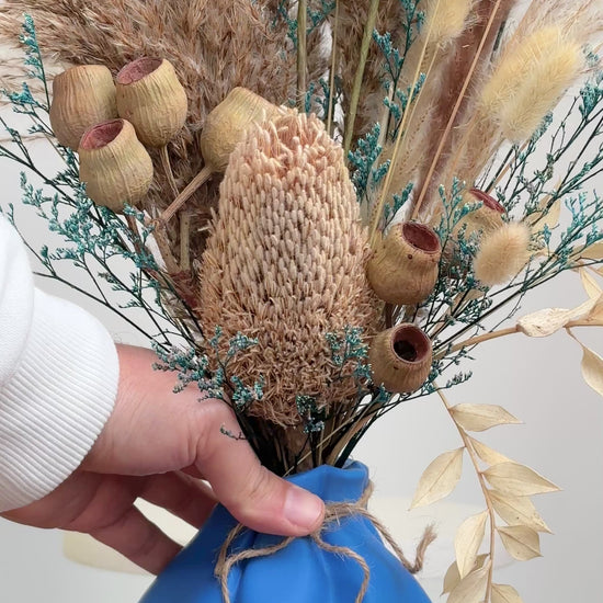Dried floral arrangement featuring pampas grass, bunny tails, and rustic seed pods in a blue vase