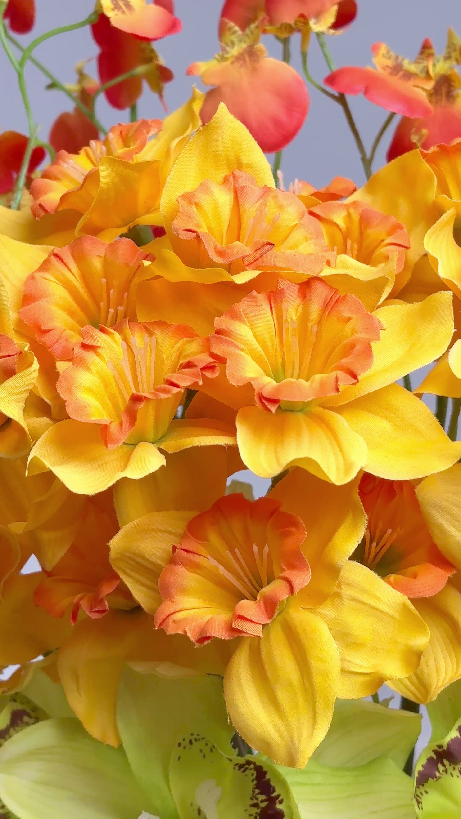 Vibrant artificial floral arrangement featuring orange orchids, yellow blossoms, and green cymbidiums in a pleated terracotta vase, styled on a white pedestal against a soft neutral background.